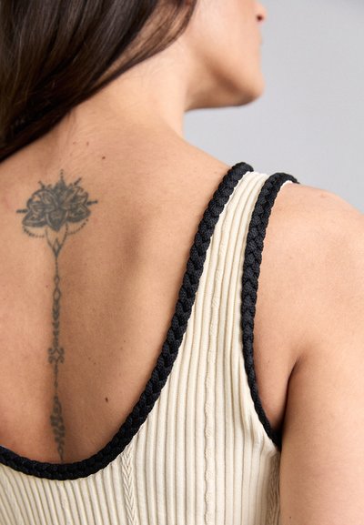 Top sin mangas con una tela texturizada en color crema y tirantes trenzados en negro. Presenta un tatuaje floral en la parte superior de la espalda.