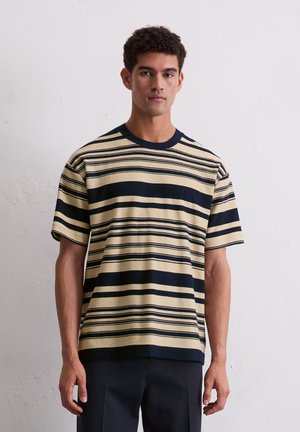 T-shirt en maille rayée avec des couleurs crème et marine, à manches courtes, col rond et tissu texturé, porté avec un pantalon marine.