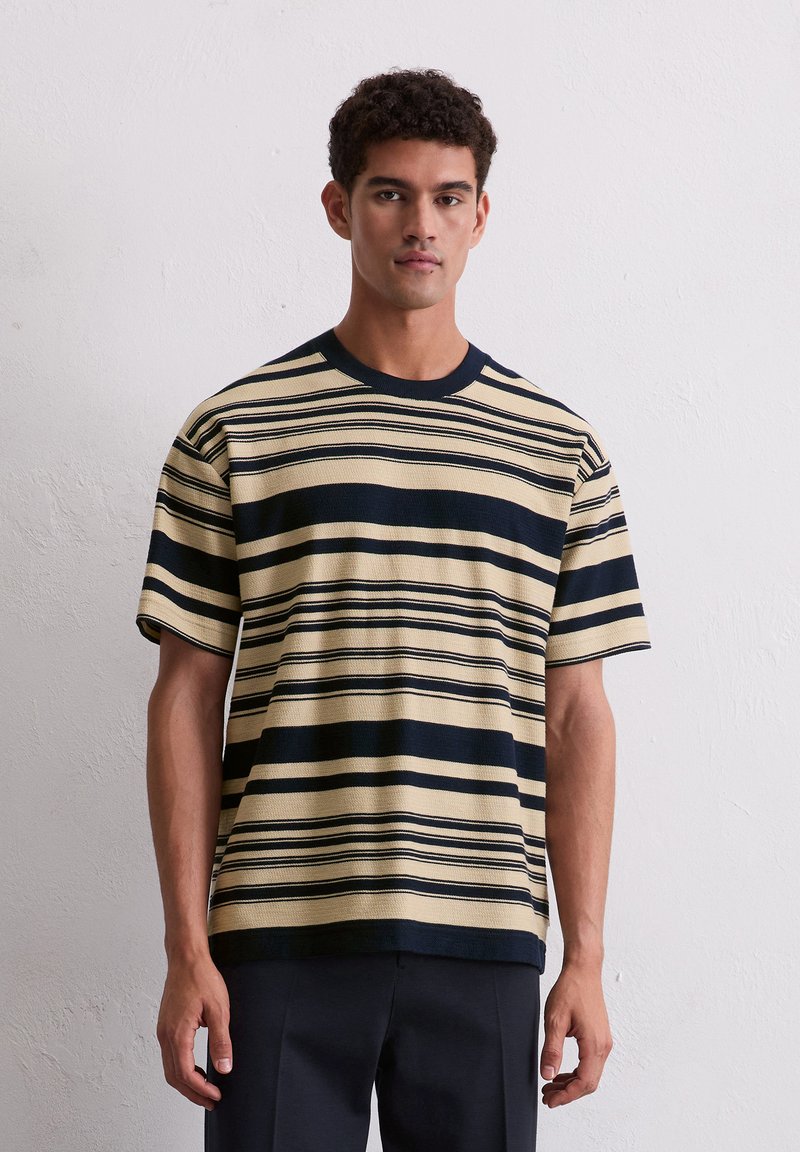 Striped knit t-shirt featuring cream and navy colors, short sleeves, crew neck, and textured fabric, worn with navy trousers.