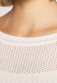 Záběr zblízka na krk a horní část hrudníku osoby, která má na sobě bílý pletený svetr se strukturovaným vzorem s otevřenými stehy.