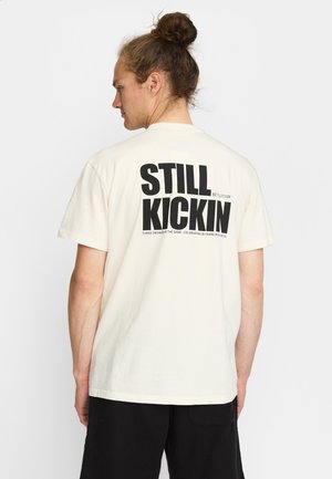 Crème katoenen t-shirt met zwarte print op de achterkant, met de woorden "STILL KICKIN" en de merknaam, korte mouwen en een ronde halsontwerp.