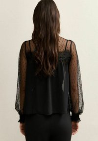 Blusa negra con mangas transparentes de lunares y detalles de encaje en los hombros, con puños fruncidos y un corte suelto.