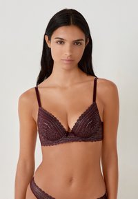 Reggiseno in pizzo bordeaux con bordi ondulati e un delicato fiocco centrale, coppe triangolari con motivo decorativo e spalline sottili regolabili.