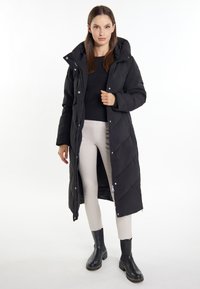 Gepolsterter schwarzer langer Mantel mit Kapuze, versehen mit einem gesteppte Muster und Druckknöpfen. Kombiniert mit hellen Hosen und schwarzen Stiefeletten.