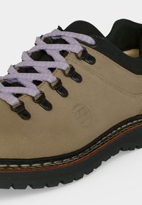 Gros plan sur une chaussure de randonnée beige avec des lacets violet clair, des œillets métalliques noirs, un logo embossé et une semelle épaisse en caoutchouc noir avec une semelle texturée.