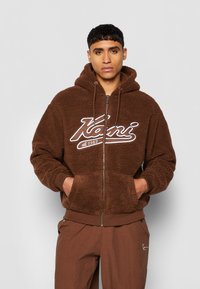 Brown zip-up hoodie with a fluffy texture, featuring a white logo on the chest and front pockets. Paired with brown pants.