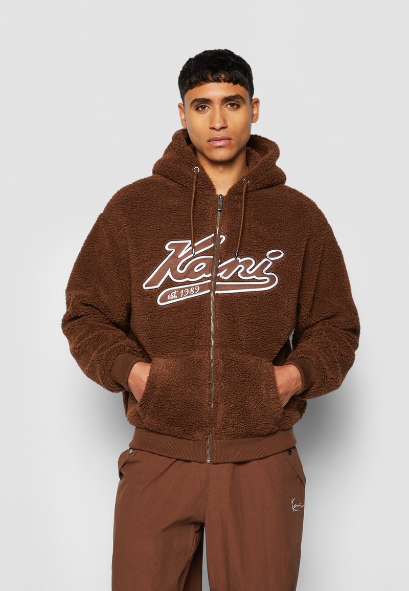 Brown zip-up hoodie with a fluffy texture, featuring a white logo on the chest and front pockets. Paired with brown pants.