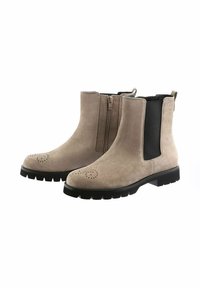 Stivaletti alla caviglia in suede beige con pannelli elastici laterali, suole nere, dettagli brogue sulla punta e una linguetta posteriore per facilitare l'indossamento.