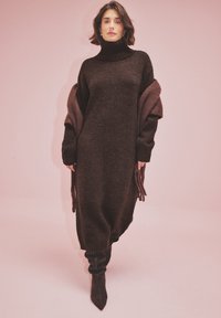 Braunes Strickkleid mit hohem Rollkragen, oversized Ärmeln und Seitenschlitzen. Kombiniert mit schwarzen, kniehohen Stiefeln. Weiche Textur und lässige Passform.