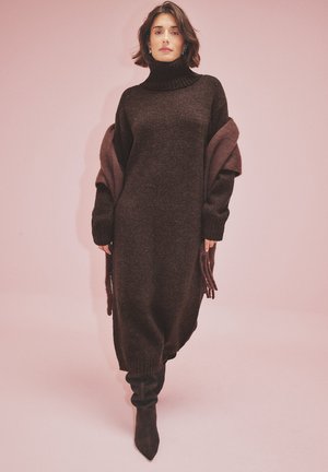 Abito in maglia marrone con collo alto, maniche oversize e spacchi laterali. Abbinato a stivali neri fino alla coscia. Tessuto morbido e vestibilità rilassata.