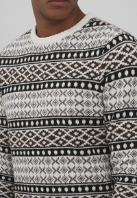 Pull en tricot à col rond, avec un motif géométrique marron, noir et blanc. Texture douce avec une apparence chaleureuse.