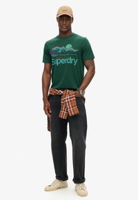 Grøn bomulds t-shirt med grafisk design, der viser bjerge og mærkenavnet "Superdry," parret med mørke jeans og en ternet flannel skjorte.