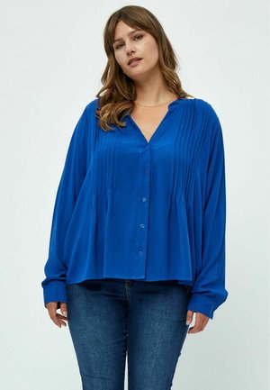Camisa azul de manga larga con botones y un diseño de frente plisado, hecha de un tejido suave. Combinada con jeans azul oscuro.