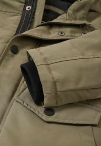 Olive green jacket featuring a zip and snap closures, with a black ribbed cuff. Smooth texture and flat construction with visible stitching.