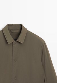 Veste vert olive avec un col pointu, des fermetures à pression et une texture de tissu lisse et légère. Coutures décoratives le long des manches.