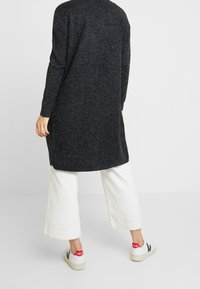 Długi czarny sweter z teksturowanej dzianiny, w zestawieniu z szerokimi białymi spodniami z kordurowej tkaniny i casualowymi białymi trampkami z czerwonymi akcentami.