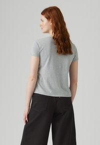 Woman with long red hair wearing a light gray short-sleeve t-shirt and black wide-leg pants, facing away against a plain background.