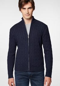 Granatowy sweter zapinany na zamek, z dzianiny w splot warkoczowy, z kołnierzem stojącym, prążkowanymi mankietami i dołem, nałożony na jasnoszary t-shirt.