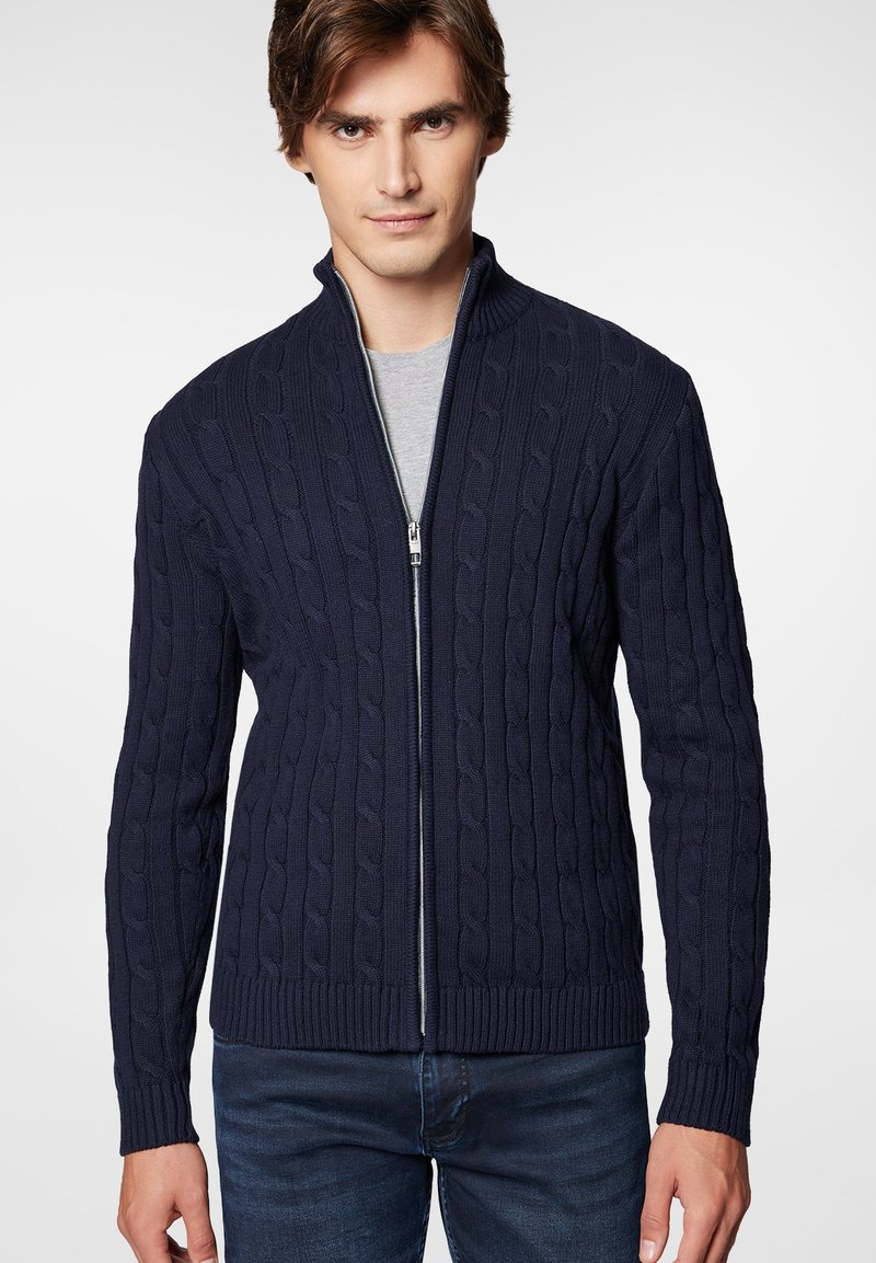 Granatowy sweter zapinany na zamek, z dzianiny w splot warkoczowy, z kołnierzem stojącym, prążkowanymi mankietami i dołem, nałożony na jasnoszary t-shirt.