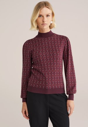 Pull en maille bordeaux avec un col montant, présentant un motif de diamant dans des couleurs contrastées. Poignets et ourlet côtelés, avec des manches légèrement bouffantes.