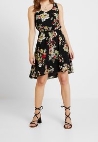 Kobieta w czarnej, bezrękawnikowej sukience w kwiaty, z przewężoną talią i czarnych sandałach na słupku, stojąca na jednolitym tle.