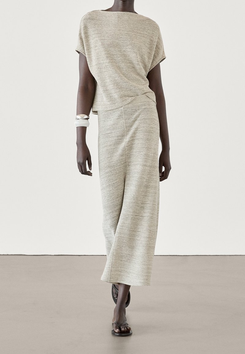 Femme portant un haut beige clair texturé à manches courtes et un pantalon large assorti, accessoirisée avec des bracelets blancs et argentés et des sandales noires.