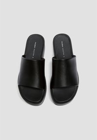 Sandales glissantes noires en cuir lisse avec une large sangle drapée et une semelle plate. Design simple sans matériel ni motifs visibles.