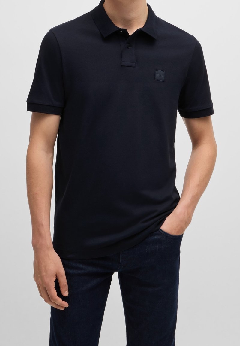 Polo bleu marine à manches courtes avec col, présentant un patch texturé sur la poitrine gauche. Porté avec un jean en denim foncé.