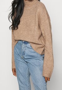 Pull en tricot beige avec un col haut, à coupe décontractée et manches longues, porté avec un jean en denim taille haute bleu clair.