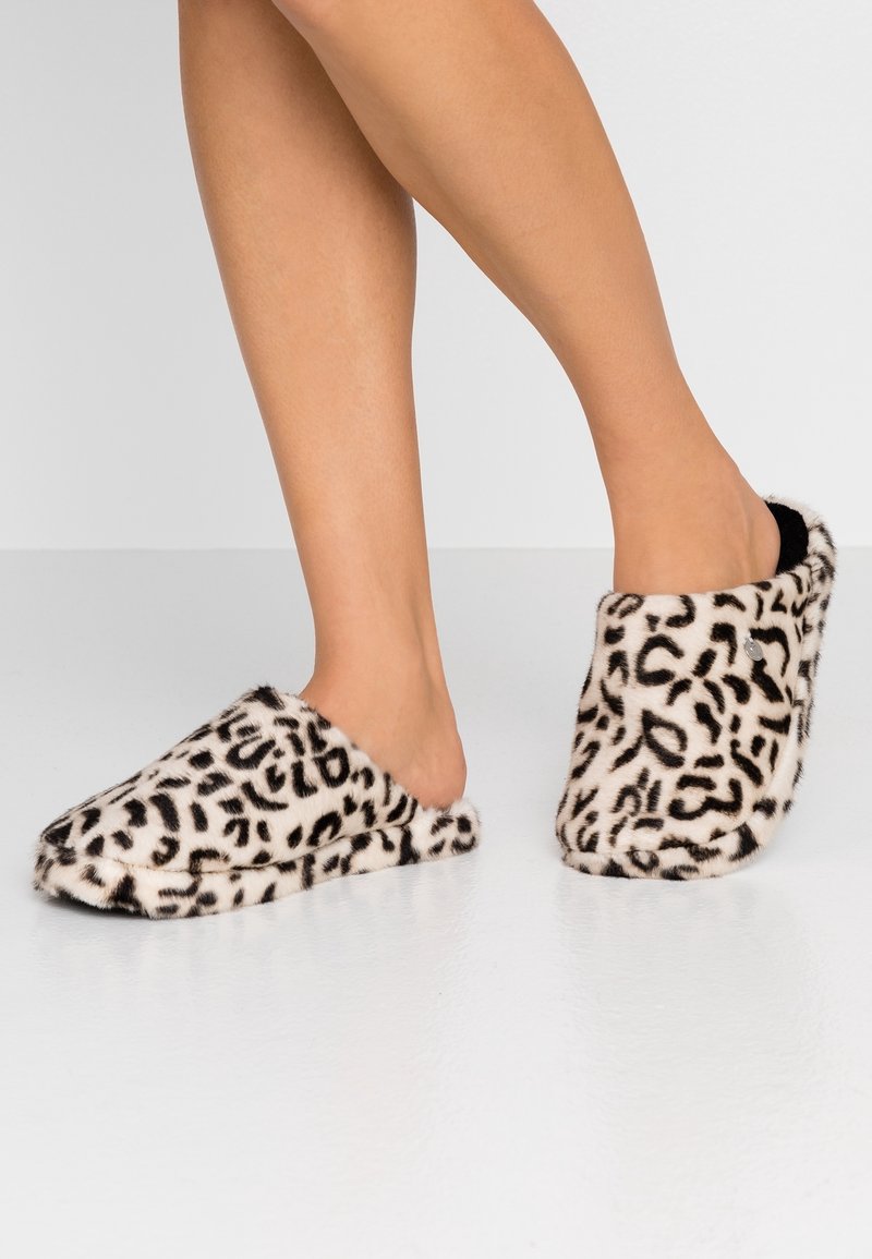 Pieds portant des chaussons en peluche faciles à enfiler avec un motif léopard blanc et noir sur un sol et un fond clairs.