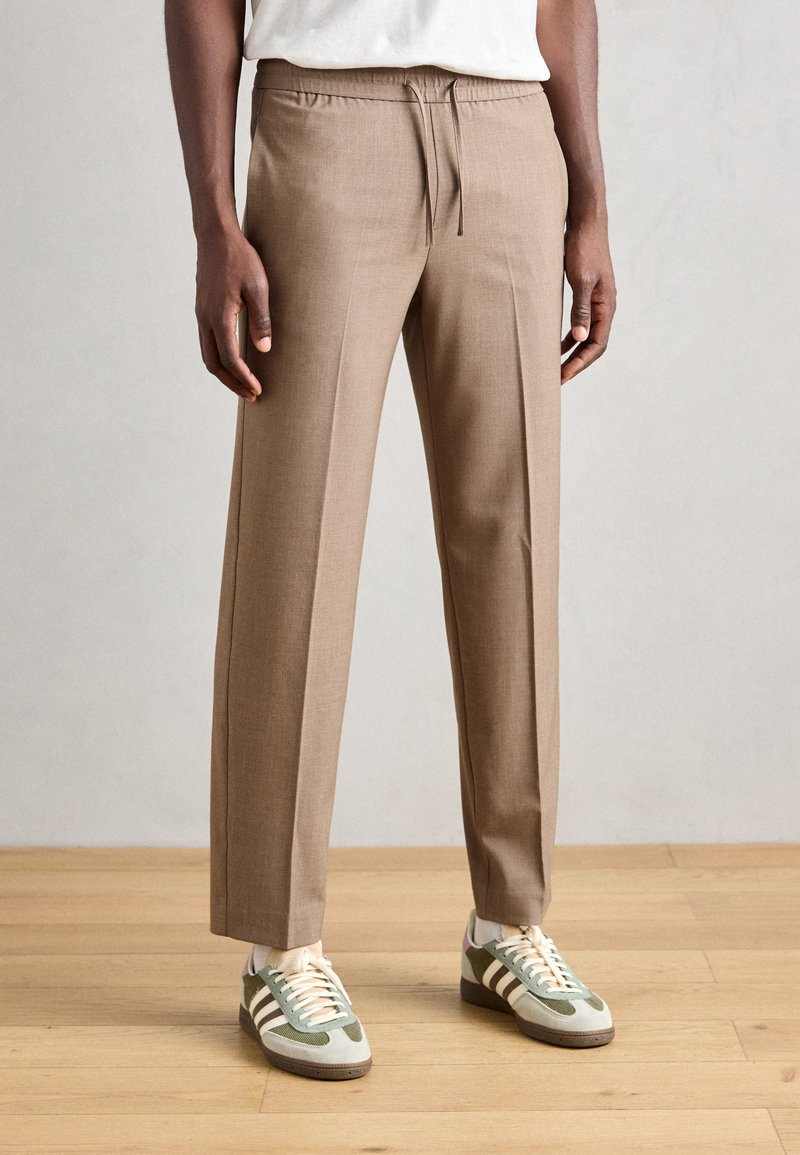 Pantalons beige ajustés avec taille élastique, jambes droites et texture lisse, associés à des baskets vertes et blanches.