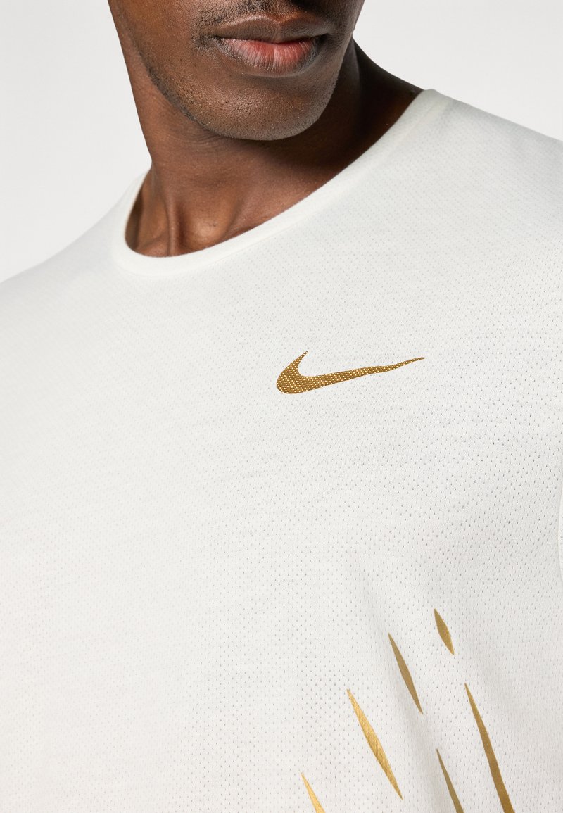 Biała, teksturowana koszulka sportowa z okrągłym dekoltem. Posiada złote logo Nike na górnym lewym boku oraz pionowe złote akcenty.