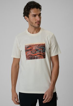 Mężczyzna o krótkich kręconych włosach, noszący beżowy t-shirt z czerwonym motywem krajobrazu i czarne spodnie, patrzy w prawo.