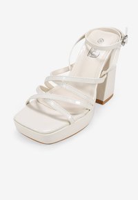 Sandalia de tacón alto de charol blanco con punta cuadrada, múltiples correas entrelazadas, hebilla en el tobillo y un tacón grueso.