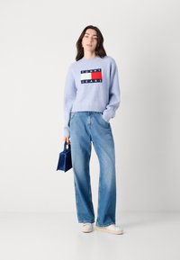 Lichtblauwe trui met geribbelde mouwen met een rechthoekig Tommy Jeans-logo. In combinatie met lose-fitting, lichtblauwe jeans en een donkerblauwe tas.