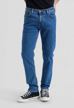 Bărbat purtând blugi denim albaștri și adidași negri cu talpă joasă și șireturi albe, stând în picioare în fața unui fundal gri deschis.