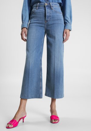 Jean bleu clair taille haute à jambes larges, porté avec des sandales à talons ouvertes rose vif et une chemise en jean bleue assortie.