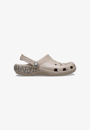 Beige klomp gemaakt van lichtgewicht materiaal met een ronde vorm. Heeft ventilatiegaten, een bandje en metalen studs op de buitenzool.