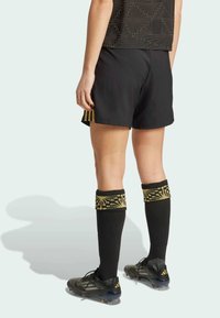 Pantaloni sportivi neri con una maglietta nera a fantasia. Calze nere con dettagli dorati, abbinate a scarpe da calcio nere e metalliche.