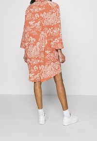 Pomarańczowa sukienka sięgająca kolan z białym tropikalnym wzorem, z szerokimi rękawami i luźnym krojem. Połączona z białymi trampkami.