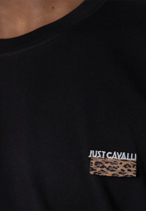 Czarny materiał z małą łatką w panterkę oraz białym, haftowanym napisem "JUST CAVALLI" w pobliżu dekoltu po lewej stronie.