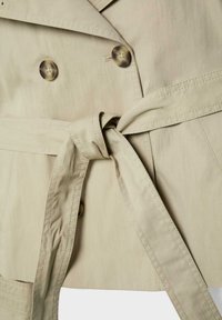 Trench-coat beige avec deux boutons marron et une ceinture en tissu nouée à la taille, présentant des coutures détaillées et un col chevauchant.