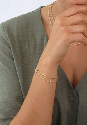 Gold-tone twisted chain bracelet with small round beads, worn on a bare arm. The top features a textured olive green fabric garment.