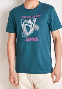 Turkusowa bawełniana koszulka z białym graficznym wzorem przedstawiającym stylizowane "CK" oraz różowym napisem "JEANS" z przodu. Krótkie rękawy, okrągły dekolt.