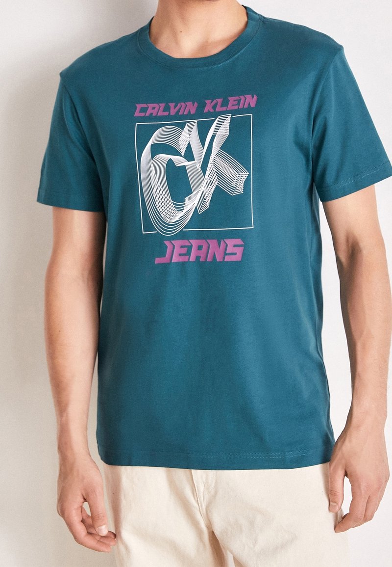 Turkusowa bawełniana koszulka z białym graficznym wzorem przedstawiającym stylizowane "CK" oraz różowym napisem "JEANS" z przodu. Krótkie rękawy, okrągły dekolt.