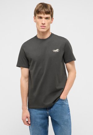 Junger Mann trägt ein dunkelgraues T-Shirt mit Mustang-Logo und blaue Jeans, steht mit einer Hand in der Tasche vor einem einfarbigen Hintergrund.