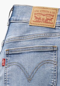 Jeans de mezclilla azul claros con una etiqueta de cuero marrón que presenta texto rojo. La distintiva costura del bolsillo trasero forma una forma de V. Etiqueta roja de Levi's visible.