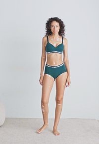 Kvinne med vitiligo iført teal sportbh og høyt midjebånd, stående barfot på teppe i nøytral rom.