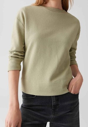Femme portant un haut texturé à manches longues de couleur olive clair avec des manches froncées et un jean noir, main dans la poche.