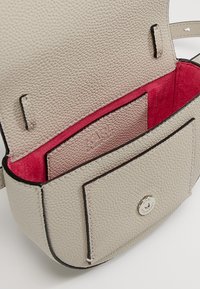 Beige, kivenmainen nahkalaukku, jossa vaaleanpunainen huopainen sisusta. Ominaisuuksiin kuuluu läppäkiinnitys, hopeiset painonapit ja sisäinen korttipaikka.